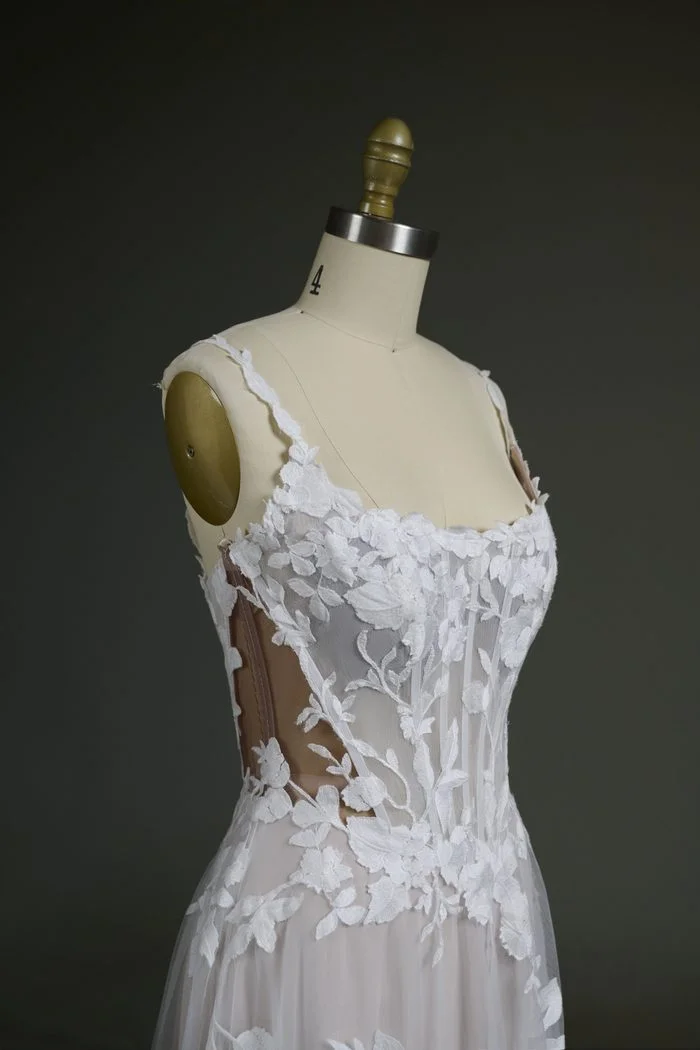 Close-up of the Natalie Marie by Maggie Sottero wedding gown bodice displayed on a dress form. The gown features a sheer corset bodice with visible boning, floral lace appliqués, and a scoop neckline. Darker nude illusion mesh is used in the side panels and bodice, demonstrating a customization option that provides a more seamless blend for deeper skin tones while preserving the gown’s transparent lace design.