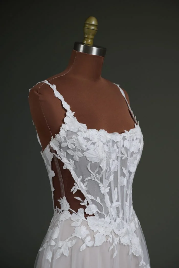 Close-up side view of the Natalie Marie by Maggie Sottero wedding gown displayed on a dress form with a dark nude mesh cover. The gown features a sheer corset bodice with exposed boning, a scoop neckline, and floral lace appliqués. The darker nude mesh highlights the illusion side panels and transparent construction, demonstrating a customization option designed to better complement deeper skin tones while maintaining the gown’s delicate lace detailing.