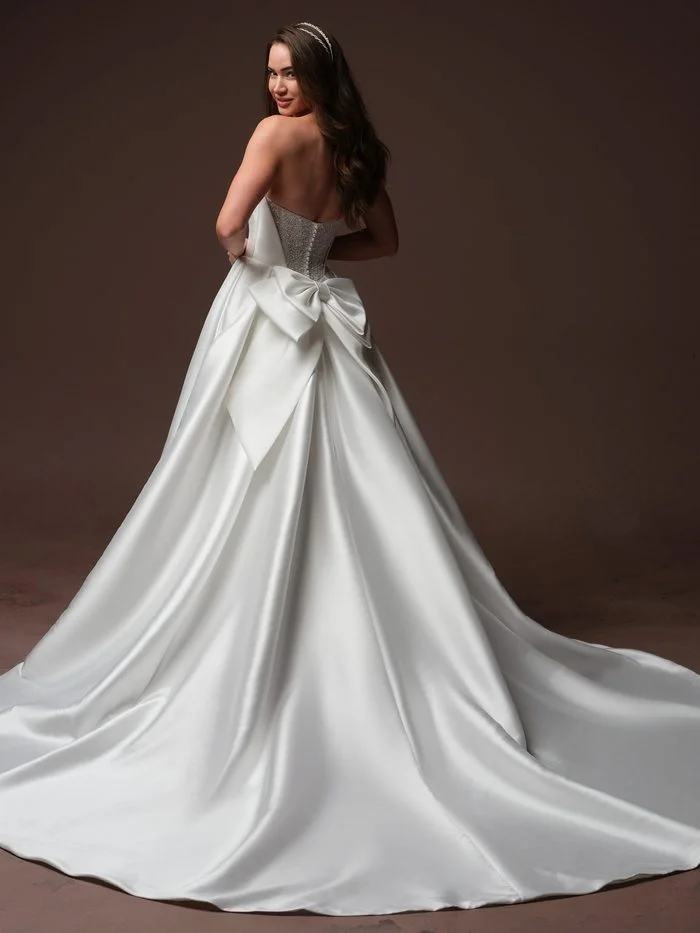 Back view of the Joanie by Rebecca Ingram wedding gown featuring a strapless satin ballgown silhouette with a low back, fitted bodice, and dramatic cathedral-length train. A statement oversized satin bow accents the back waist, creating a striking focal point while adding volume and romance to the embelished design.