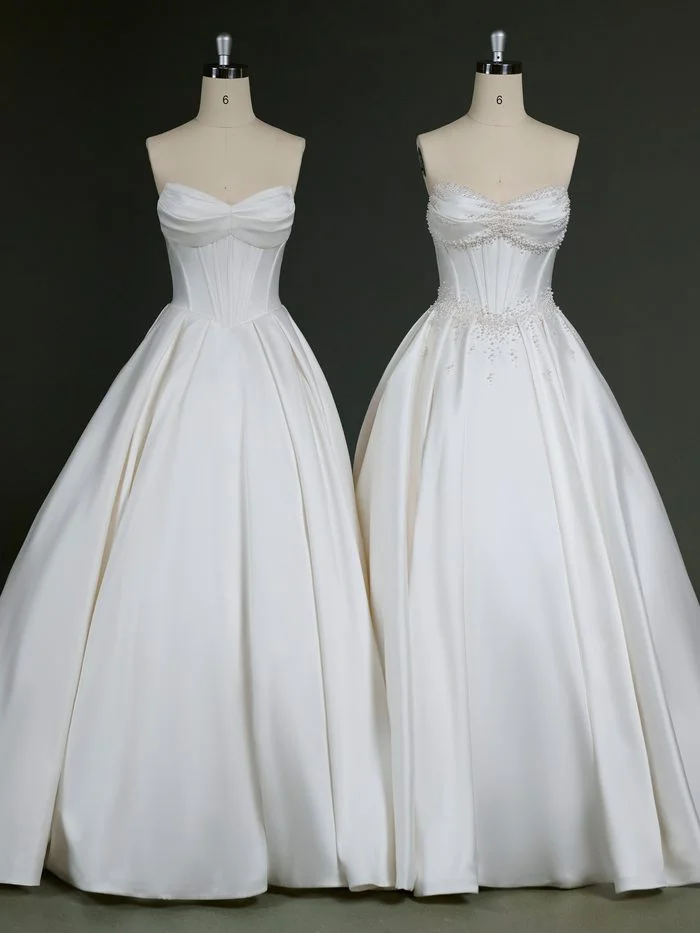 Two versions of the Juneau by Maggie Sottero wedding gown are displayed side by side on dress forms. Both feature a strapless satin ballgown silhouette with a draped sweetheart neckline, corset-inspired bodice, and full pleated skirt. The gown on the left showcases a clean, minimalist design, while the gown on the right is customized with pearl embellishments along the neckline, waist, and upper skirt, adding dimensional sparkle to the classic silhouette.