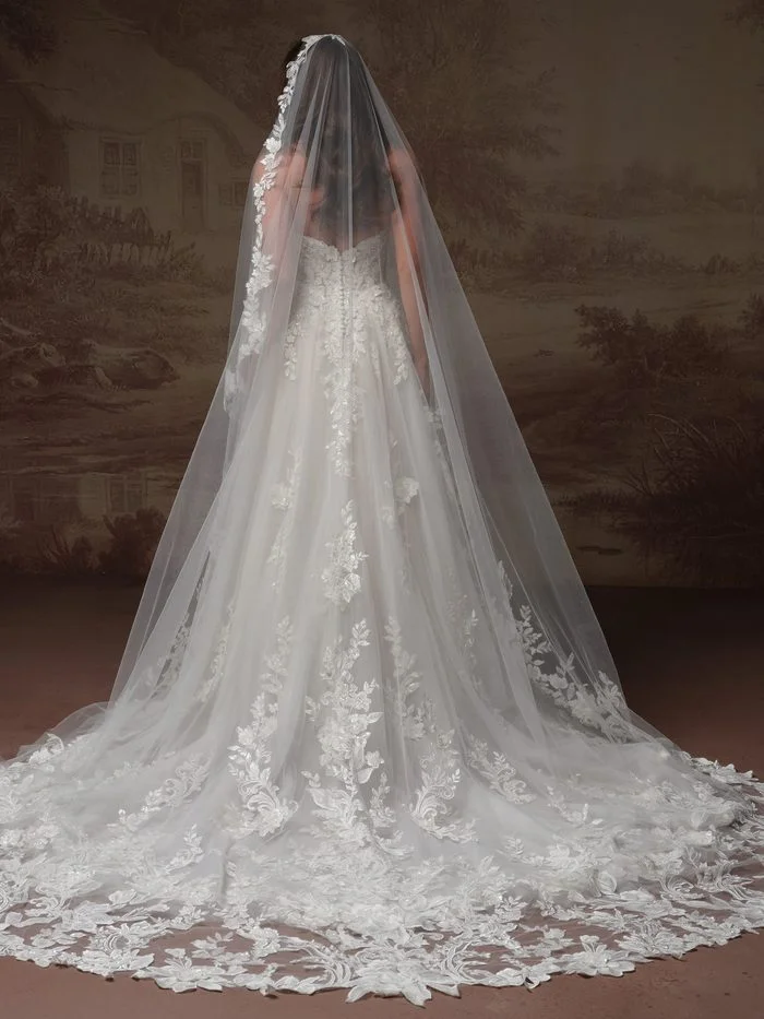 Back view of the Bellevue Cape accessory by Maggie Sottero, featuring a dramatic cathedral-length cape crafted from delicate floral lace. The cape includes an attached lace hood and cascades from the shoulders into a sweeping train, showcasing intricate lace motifs and scalloped edges throughout the design.