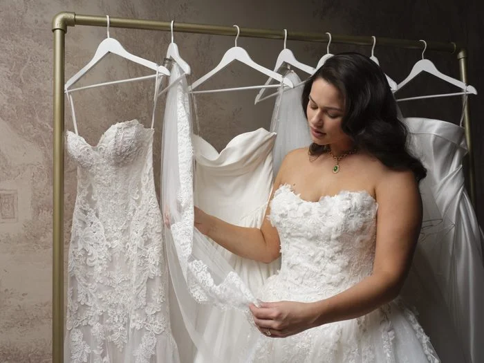 Lifestyle image featuring the Benton by Maggie Sottero wedding gown among a collection of bridal styles on a garment rack. In the foreground, the Benton gown showcases a strapless sweetheart neckline and dimensional floral appliqués that create texture across the bodice and skirt. A coordinating lace-trimmed veil is draped through the hands, while additional wedding gowns with lace, tulle, and satin details hang in the background, highlighting bridal styling and accessory options.