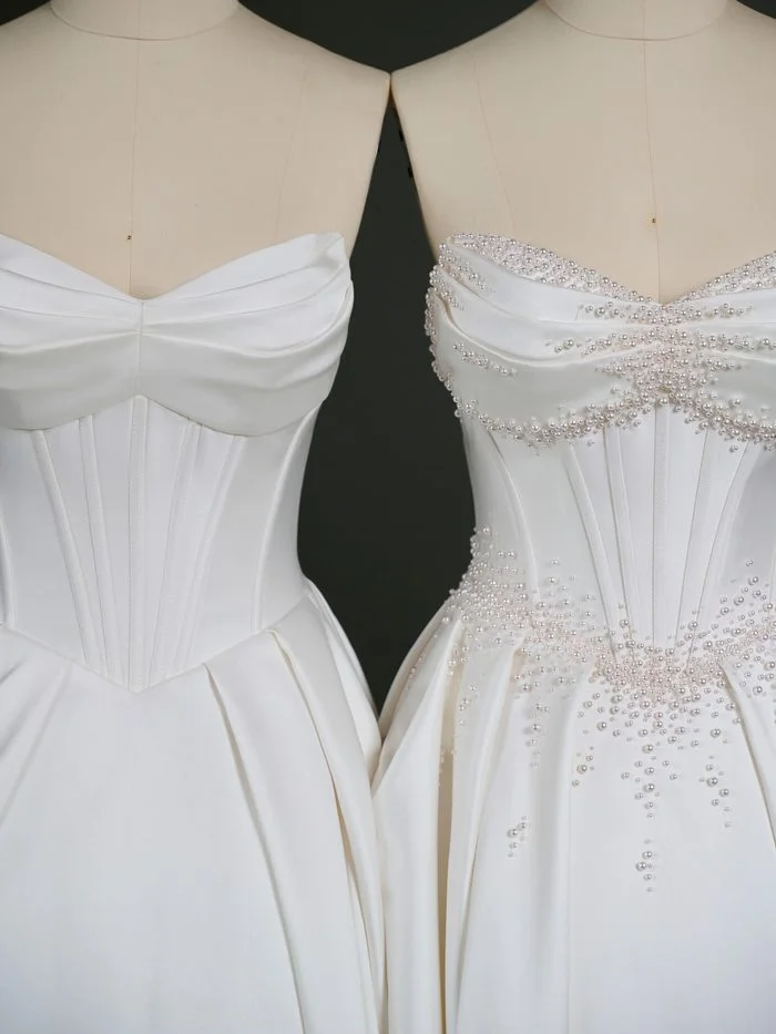 Close-up comparison of two Juneau by Maggie Sottero wedding gown bodices. The left version features a clean satin corset bodice with a softly draped sweetheart neckline and visible boning channels. The right version showcases the same satin bodice enhanced with hand-applied pearl beading concentrated along the neckline, center front, waist, and upper skirt, highlighting a customizable embellished finish.