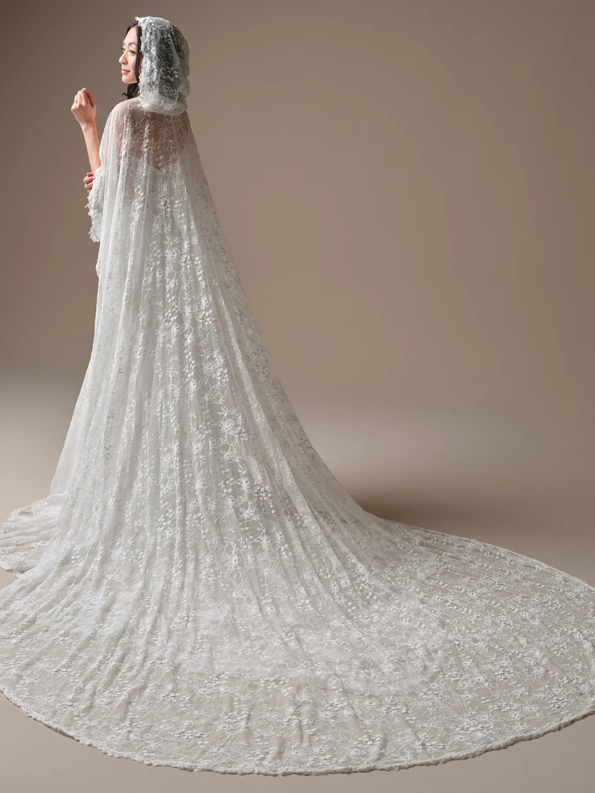 Back view of the Bellevue Cape accessory by Maggie Sottero, featuring a dramatic cathedral-length cape crafted from delicate floral lace. The cape includes an attached lace hood and cascades from the shoulders into a sweeping train, showcasing intricate lace motifs and scalloped edges throughout the design.