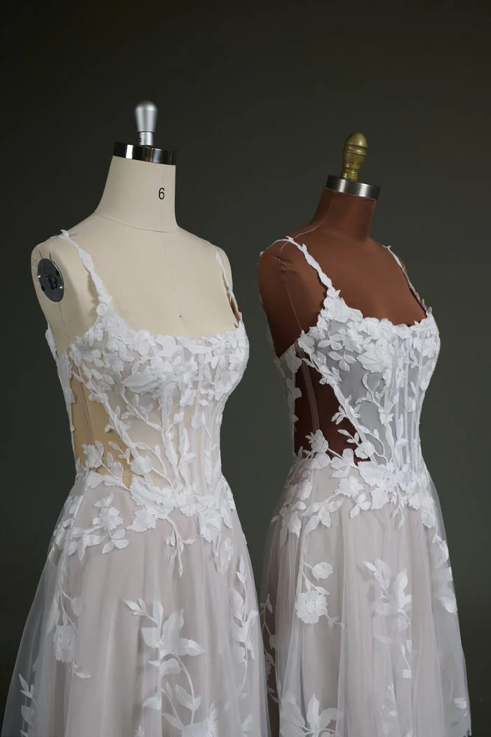 Two versions of the Natalie Marie by Maggie Sottero wedding gown are displayed side by side on dress forms, demonstrating different illusion mesh color options. Both feature a sheer corset bodice with exposed boning, a scoop neckline, floral lace appliqués, and a soft tulle skirt. The gown on the left showcases a lighter nude illusion mesh, while the gown on the right features a darker nude illusion mesh designed to better complement deeper skin tones while maintaining the gown’s delicate sheer effect.