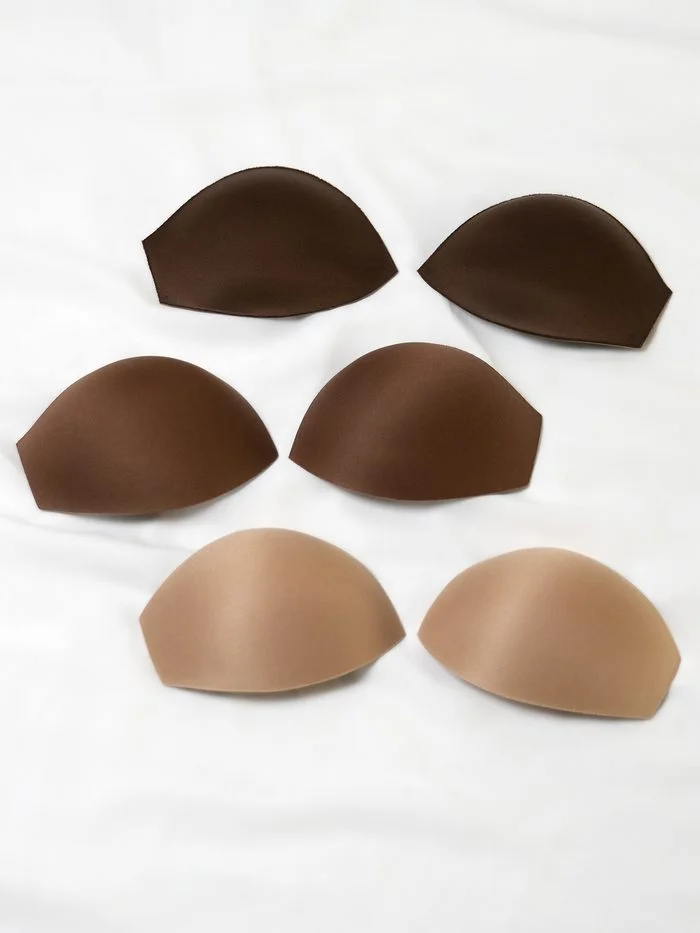 Six bridal bra cups by Maggie Sottero displayed on a white fabric background. The molded cups are shown in three inclusive nude shades ranging from light to dark, arranged in matching pairs to illustrate available color options for added support and coverage in wedding gowns.