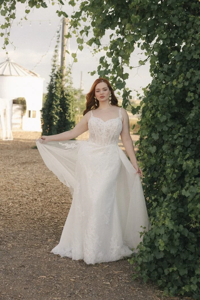 Full-length view of the Esmeralda Lane boho floral wedding dress with cascading floral lace, fitted silhouette, and soft tulle overskirt framed by lush green vines.