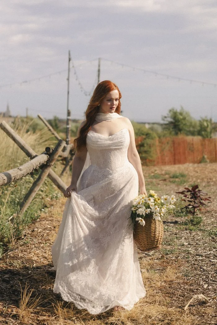Maelle boho floral wedding dress featuring allover lace, a draped sweetheart neckline, and a soft matching neck scarf, styled in a romantic countryside setting with wildflowers.