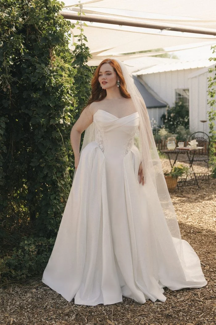 Gigi boho-inspired wedding dress by Rebecca Ingram featuring a draped strapless neckline, lace-accented corset bodice, and a full satin ballgown skirt with pockets in a romantic garden setting.