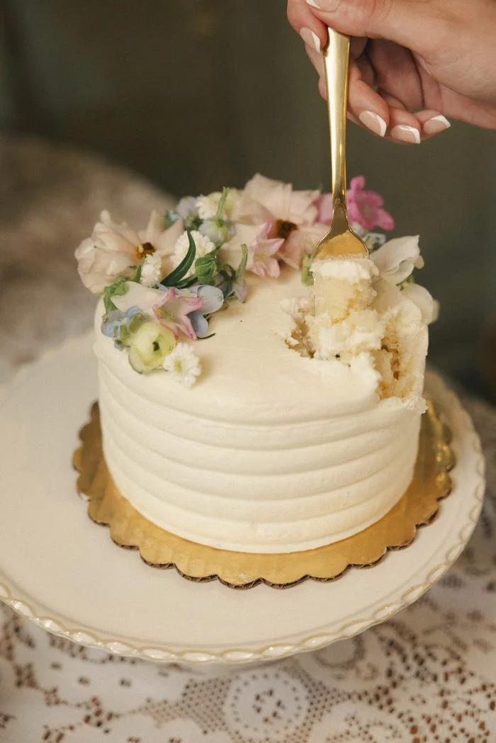 Small white wedding cake with smooth buttercream frosting and fresh pastel flowers, styled on a vintage lace-covered table as part of a romantic boho wedding dessert display.