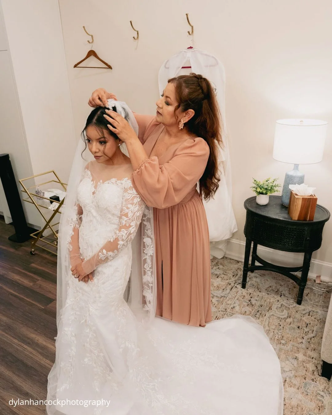 a bride in a white lace gown sits on a plush rug as her mother gently adjusts her veil dylanhancockphotography