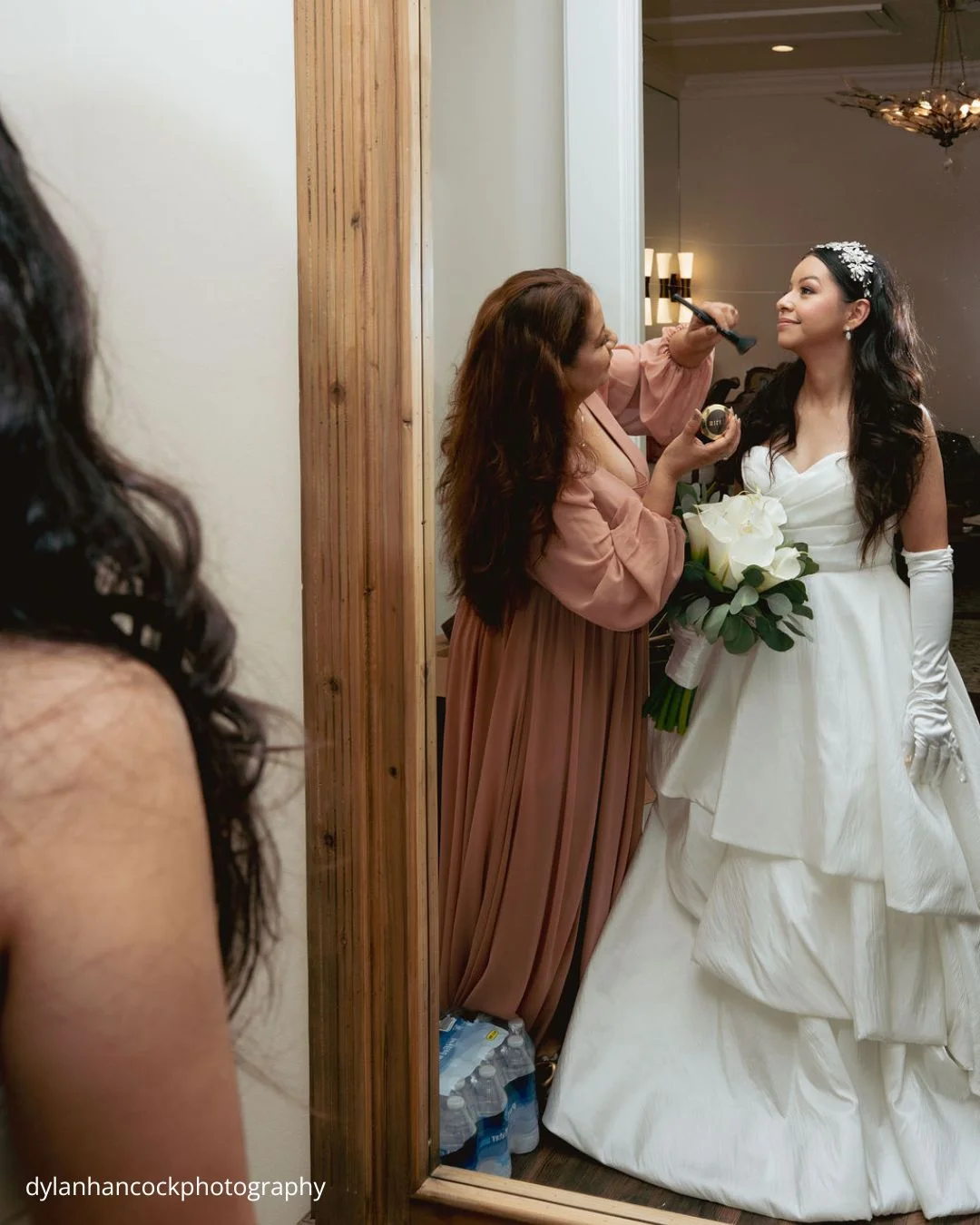 a bride in a white gown and gloves stands before a large wooden framed mirror as a mom beside her applies makeup dylanhancockphotography