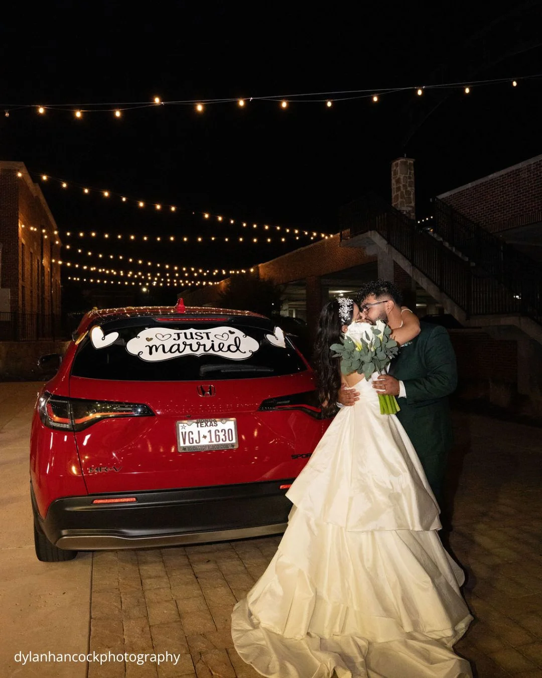 a couple shares an intimate kiss in front of their Just Married decorated car under the warm glow of string lights at night dylanhancockphotography