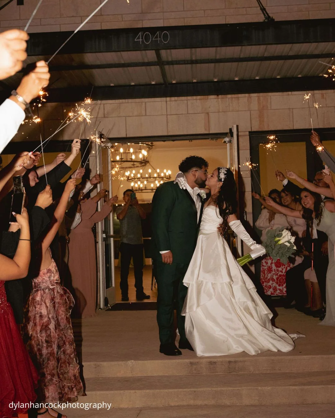 a couple shares a kiss as they exit a building surrounded by guests holding sparklers in celebration dylanhancockphotography