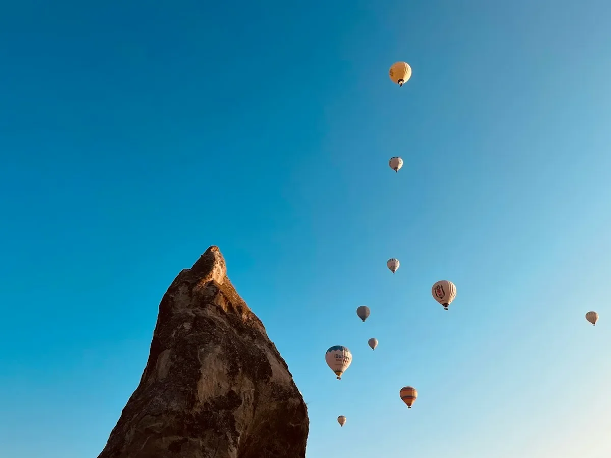 Colorful hot air balloons rising past a tall rock spire against a clear blue sky in Cappadocia