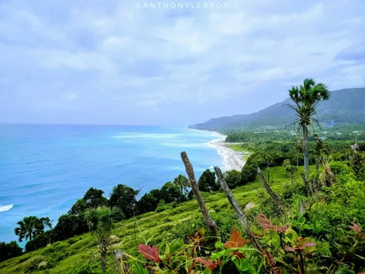 A breathtaking coastal view of lush green cliffs overlooking the turquoise waters of the ocean, with a secluded beach stretching along the shoreline under a partly cloudy sky.