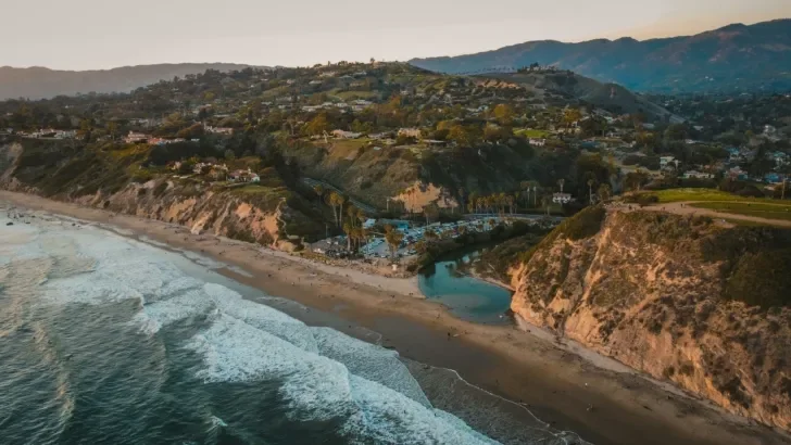 A breathtaking aerial view of a coastal landscape at sunset, showcasing rugged cliffs, a sandy beach, and ocean waves rolling onto the shore, with palm trees and lush green hills in the background.
