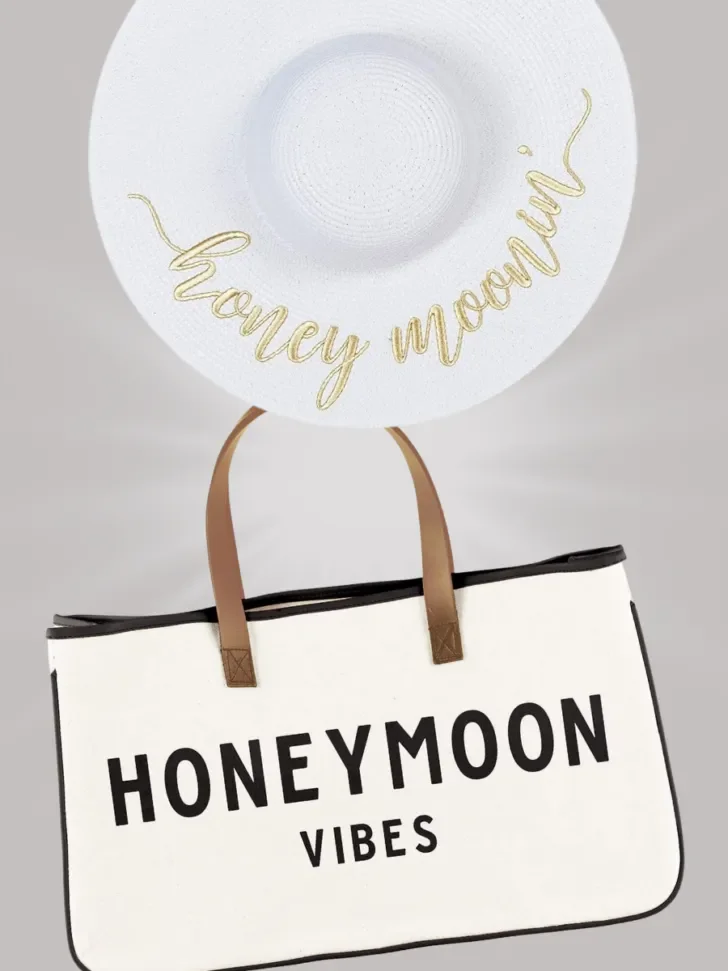White wide-brimmed hat with "Honeymoonin'" written in gold and a canvas tote bag with "Honeymoon Vibes" written in bold black letters.